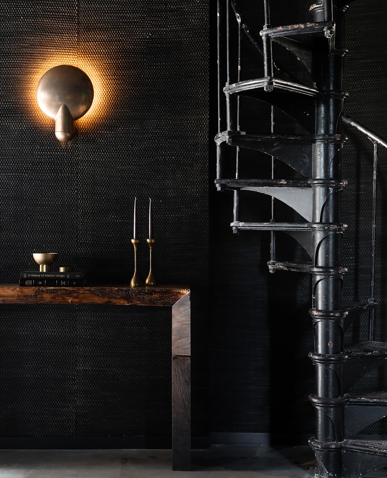 Black textured wall with a glowing brass sconce, live-edge console and cast-iron spiral staircase