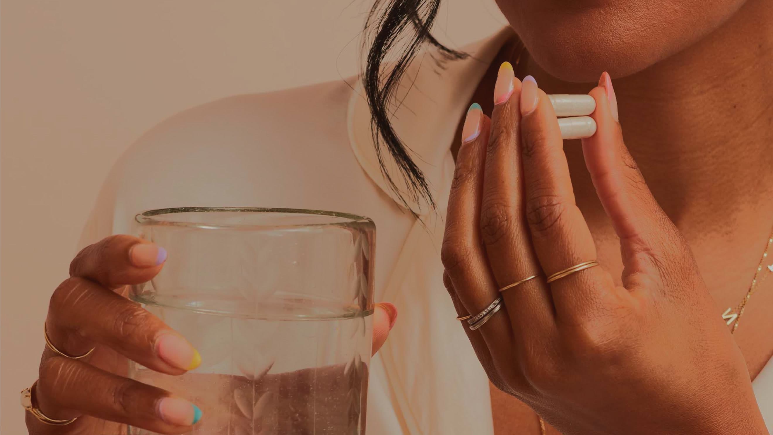 Full-width warm-toned lifestyle photo, close crop of a woman holding a glass of water in one hand and two capsules to her collarbone in t…