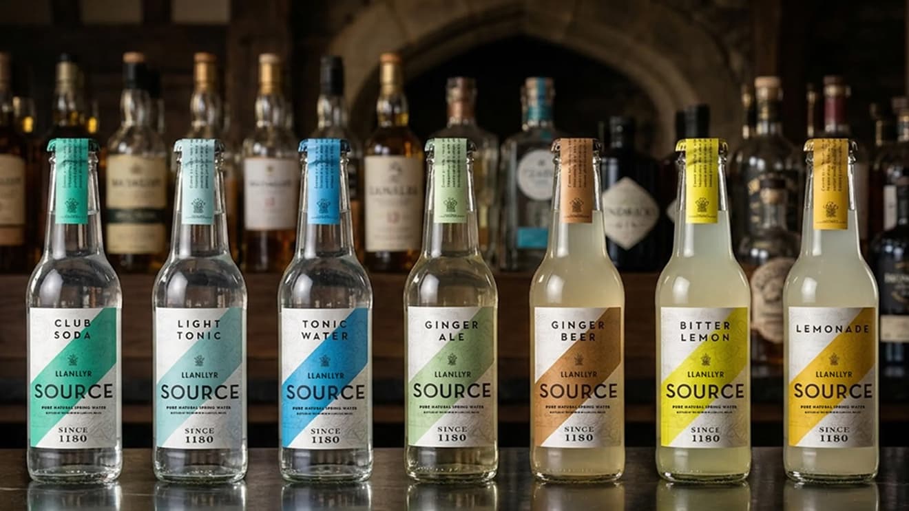 Full-width photo of the Llanllyr Source mixer range lined up on a bar top in front of a back bar of spirits, with reflections below