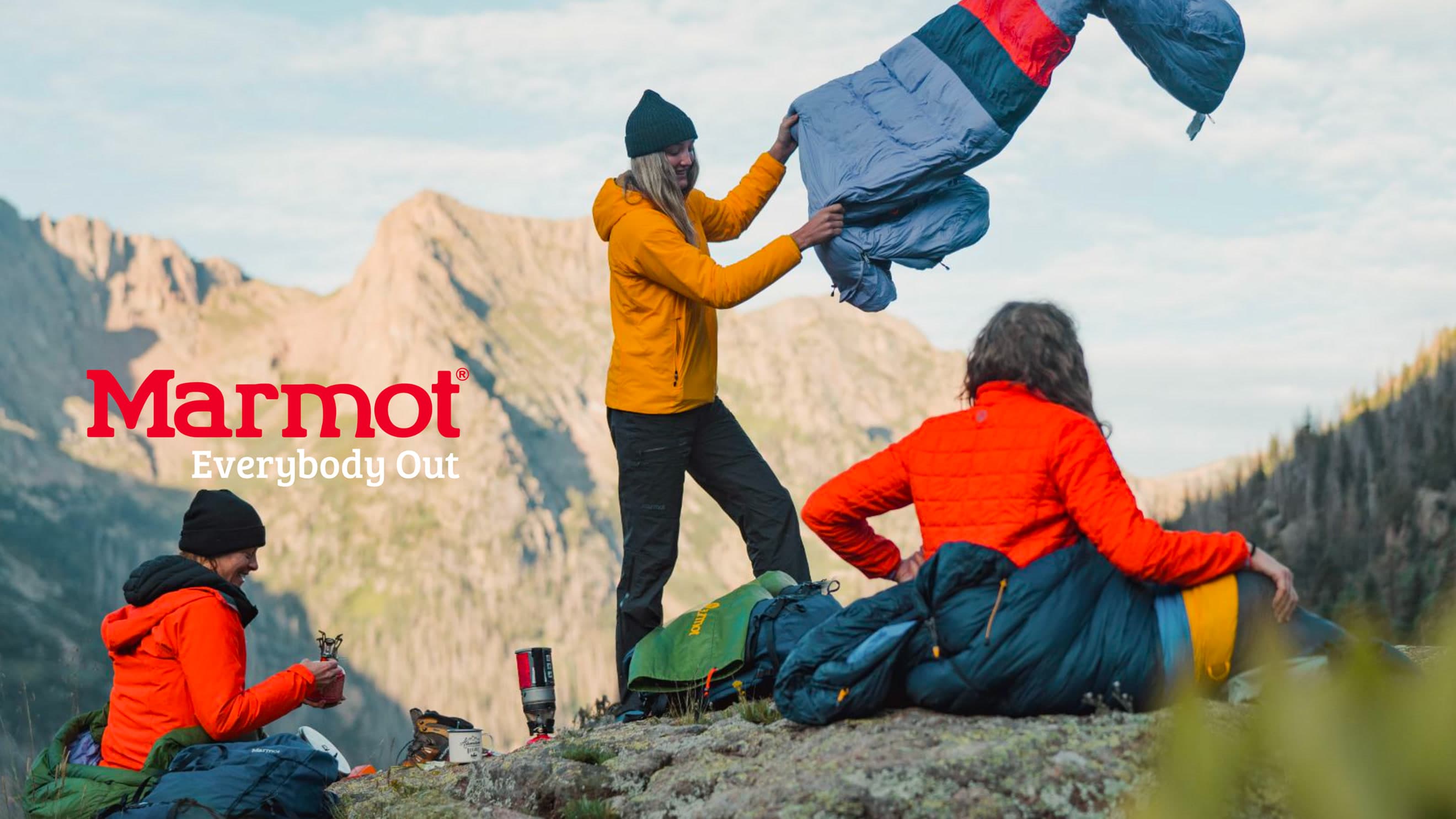 Wide campaign photo: a woman in a mustard jacket shaking out a blue/red sleeping bag on a mountain ridge, friends in orange jackets seate…