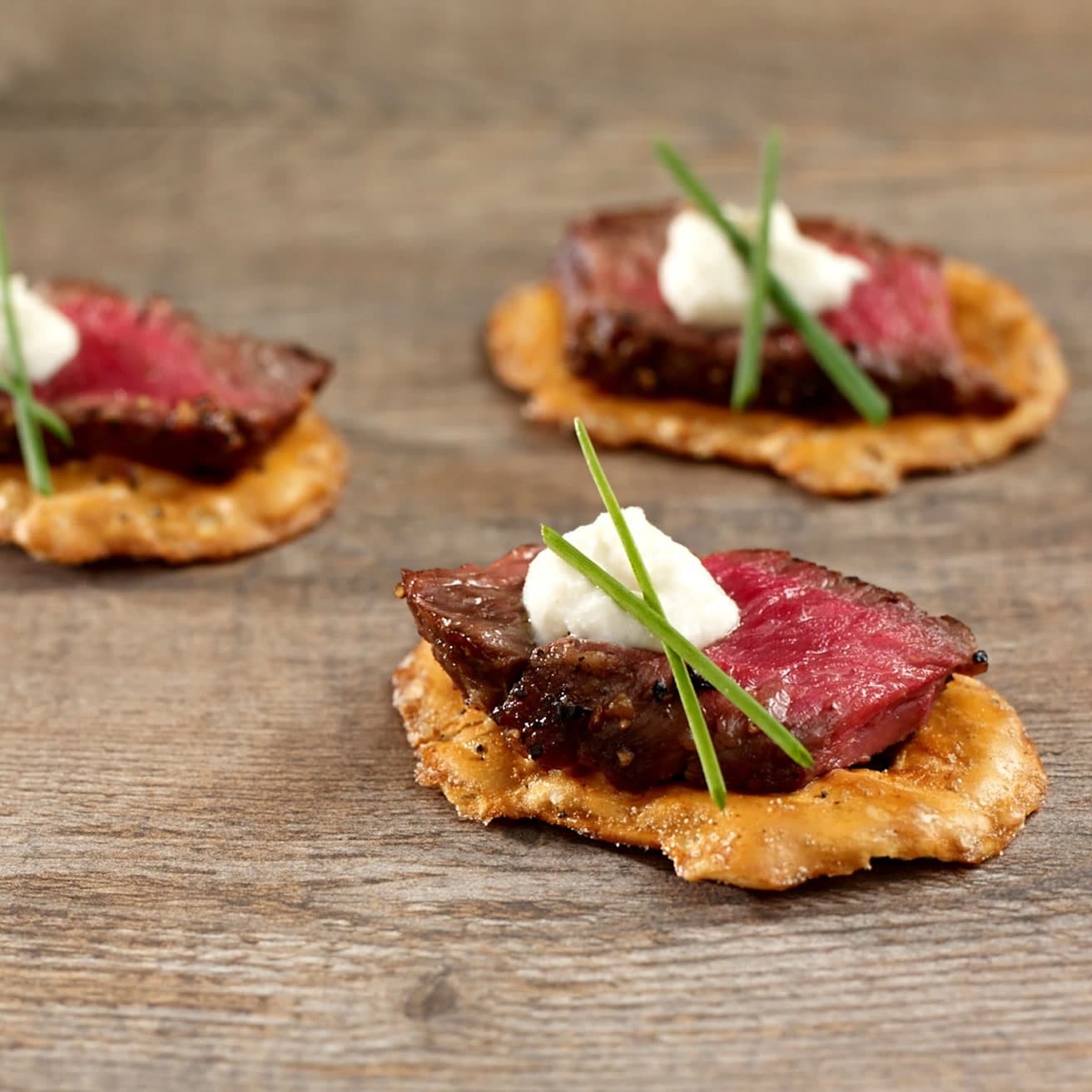 Left: close-up food photo of Pretzel Crisps topped with seared rare steak, horseradish cream and chives on weathered wood. (1 of 2)