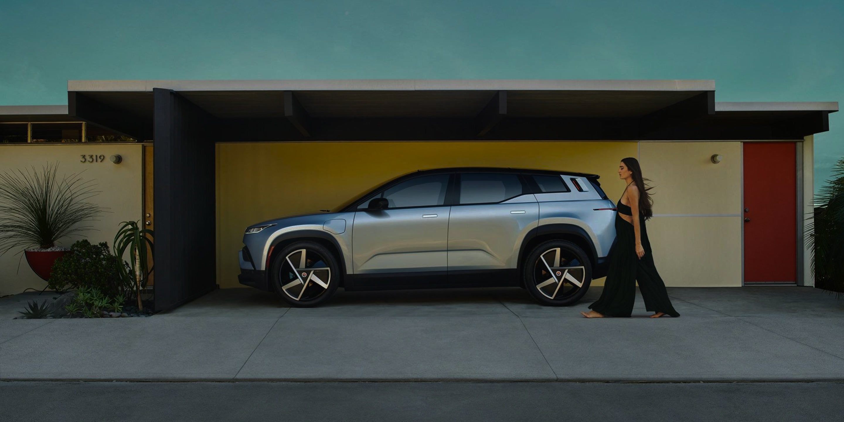 full-bleed photo of a silver Fisker Ocean parked under the carport of a mid-century modern house at dusk, woman in a black dress walking…