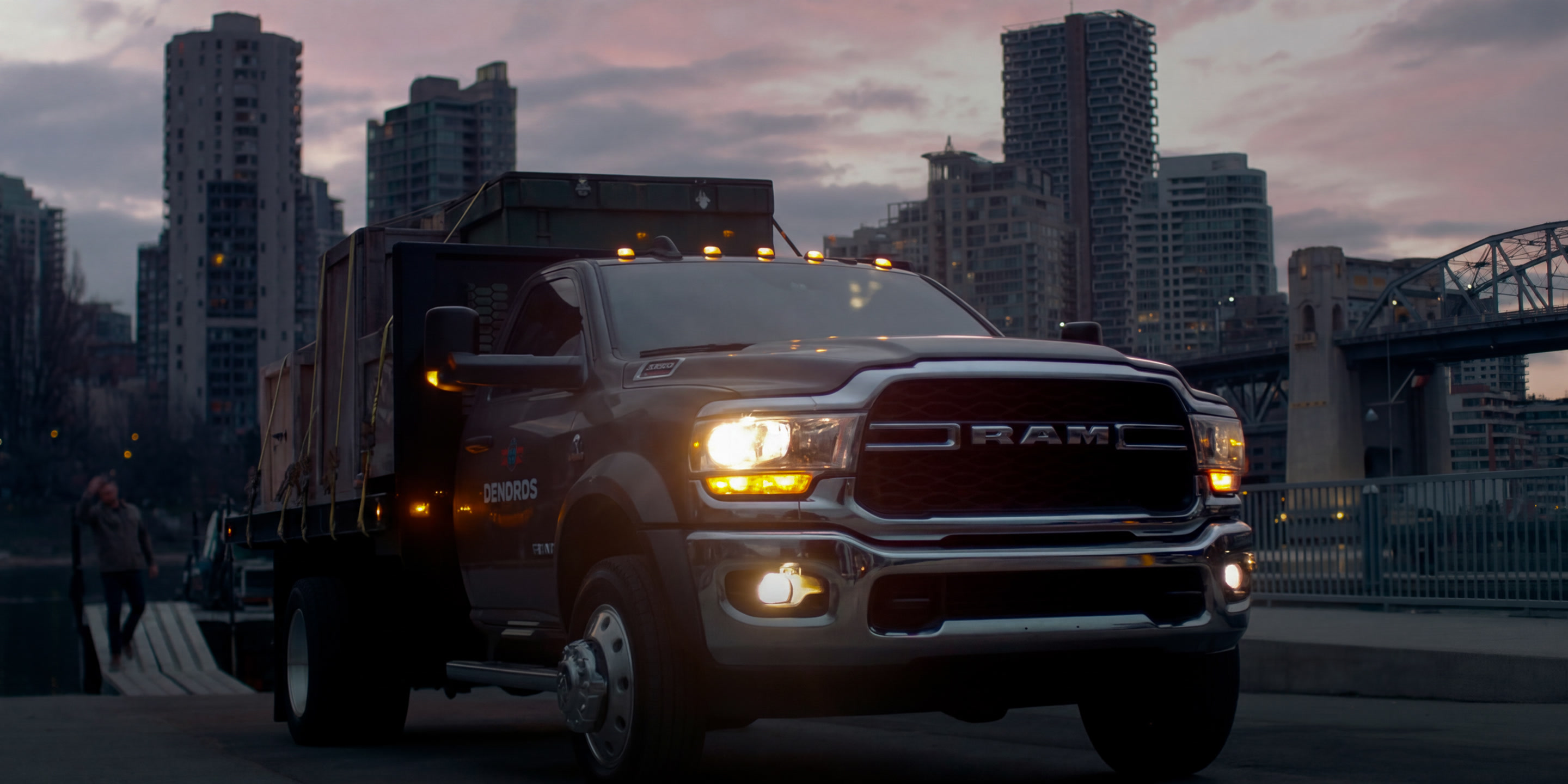 full-bleed photo of a Ram 5500 dump truck at dusk in front of a city skyline and bridge; headline set in white over the lower left